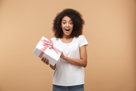Portrait of an excited young african woman holding present box isolated over beige backgroundの写真素材