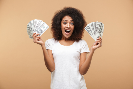 Portrait of a happy young african woman holding bunch of money banknotes isolated over beige backgroundの写真素材