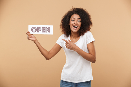 Portrait of an excited young african woman pointing finger at a sign OPEN isolated over beige backgroundの写真素材