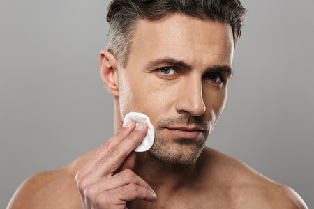 Image of serious concentrated mature man standing isolated over grey wall background take care of his skin.の写真素材
