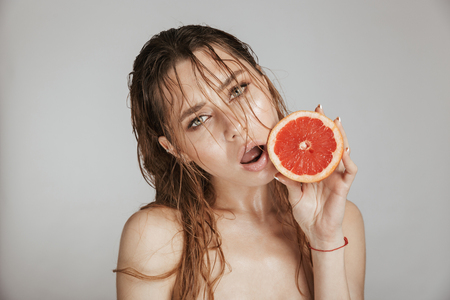 Fashion portrait of a topless beautiful woman with makeup and wet hair holding sliced grapefruit isolated over gray backgroundの写真素材