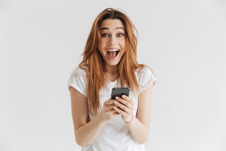 Surprised happy woman in casual clothes holding smartphone and looking at the camera over grey backgroundの写真素材
