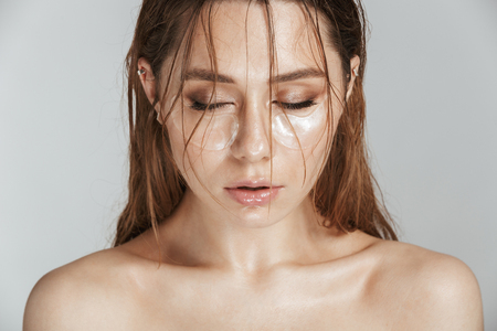 Close up fashion portrait of a topless beautiful woman with makeup and wet hair wearing cosmetic eye patches isolated over gray backgroundの写真素材