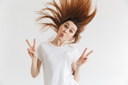 Funny woman in t-shirt having fun while showing peace gestures and looking at the camera over grey backgroundの写真素材