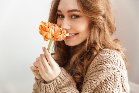 Photo of lovely woman in sweater sitting at table in cafe looking on camera with smile and smelling flowerの写真素材
