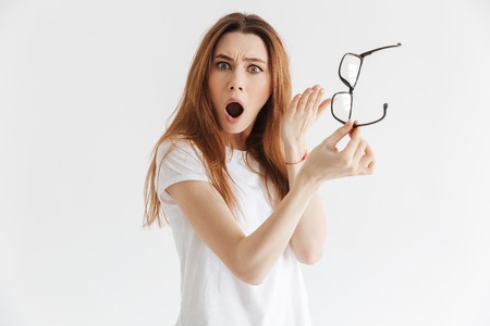 Shocked woman in t-shirt showing eyeglasses and looking at the camera over grey background