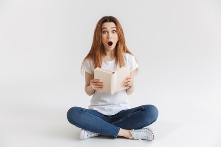 Shocked woman in t-shirt sitting on the floor while holding book and looking at the camera over grey backgroundの写真素材
