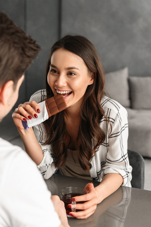 Photo of lovely smiling woman looking at man while sitting together at table in living room and eating chocolate with teaの写真素材