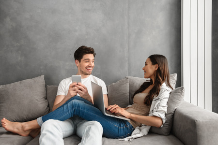 Photo of loving couple man and woman 30s lying in living room on gray sofa and smiling while using modern technicsの写真素材