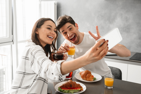 Portait of beautiful family happy man and woman taking selfie on smartphone while having breakfast indoorの写真素材