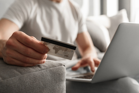 Photo cropped of caucasian guy 30s in casual jeans using notebook and holding credit card for online shopping while sitting on sofa at homeの写真素材