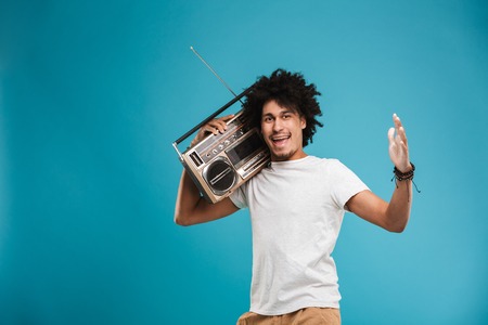 Image of excited young african curly man standing isolated over blue background holding boombox.の写真素材