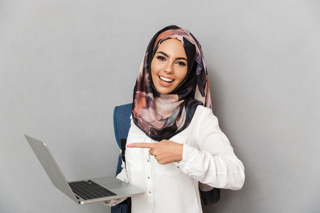 Portrait of a cheerful young arabian woman student with backpack pointing finger at laptop computer isolated over gray backgroundの写真素材
