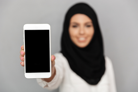 Blurry image of smiling muslim woman 20s in islamic headscarf with oriental makeup demonstrating closeup cell phone at camera isolated over gray backgroundの写真素材