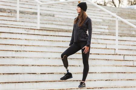 Photo of healthy disabled woman in sportswear with prosthesis leg standing at the stairs and looking aside outdoorの写真素材