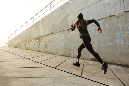 Portrait of disabled athlete woman with prosthetic leg in tracksuit running along concrete wall outside