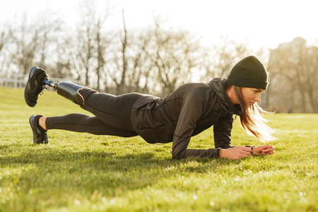 Image of sporty handicapped woman in black tracksuit warming up and doing plank with prosthesis leg on natureの写真素材