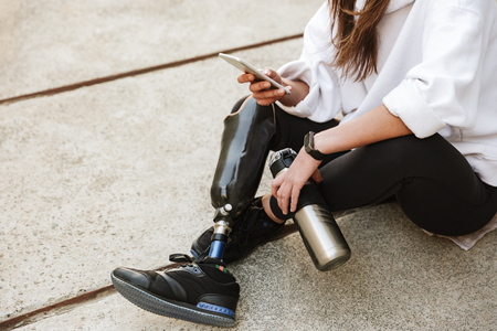 Cropped photo of handicapped woman in streetwear having bionic leg sitting on concrete floor outdoor and using cell phoneの写真素材