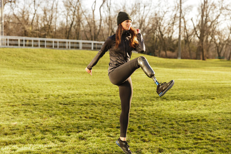 Image of happy handicapped woman 20s in tracksuit doing sports on nature and raising prosthesis legの写真素材