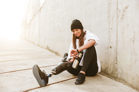 Photo of content handicapped woman having bionic leg in streetwear sitting on concrete floor outdoor and looking at wristwatch with cup in handの写真素材