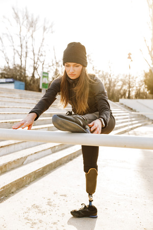 Image of disabled running girl in sportswear training and stretching prosthetic leg on railing in stadium outsideの写真素材