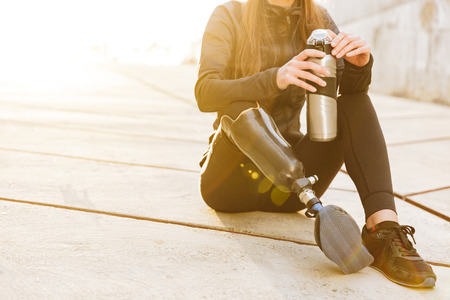 Cropped photo of athletic disabled girl with prosthetic leg in sportswear sitting on concrete floor outdoors and holding  cupの写真素材
