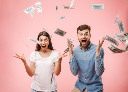 Portrait of a joyful young couple standing under money banknotes shower and celebrating success isolated over pink backgroundの写真素材