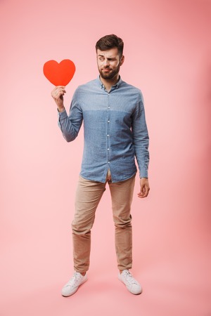 Full length of a confused young man holding red heart isolated over pink backgroundの写真素材
