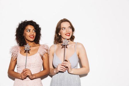 Portrait of two pretty well dressed women having fun with magical wands isolated over white backgroundの写真素材