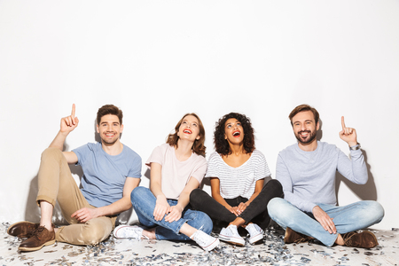 Group of happy multiracial people sitting on a floor with confetti and pointing up isolated over white backgroundの写真素材