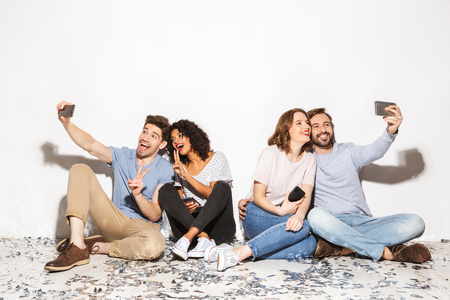 Group of happy multiracial people sitting on a floor with confetti and taking selfies isolated over white backgroundの写真素材