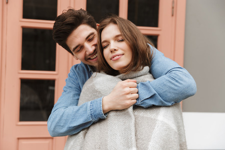 Photo of happy couple in love standing outside cafe while man hugging and wrapping his woman in warm blanketの写真素材
