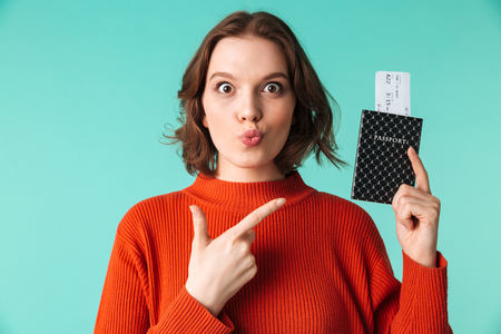 Portrait of a cute young woman dressed in sweater pointing finger at a passport with flying tickets isolated over blue backgroundの写真素材