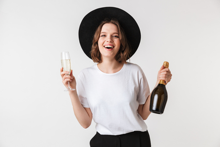 Portrait of a cheerful young woman dressed in black hat holding bottle of champagne and a glass isolated over white backgroundの写真素材