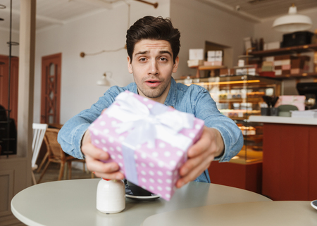 Photo of handsome man giving gift box at camera while sitting at table in cafe or bakeryの写真素材