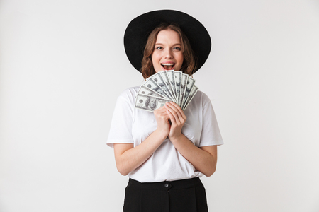Portrait of an excited young woman dressed in black hat holding money banknotes isolated over white backgroundの写真素材
