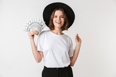 Portrait of a cheerful young woman dressed in black hat holding money banknotes isolated over white backgroundの写真素材