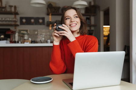 Image of happy caucasian woman 20s smiling while using laptop and drinking coffee in cafeの写真素材