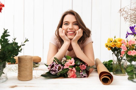 Photo of cute cheerful florist woman standing near table working with flowers in workshop. Looking camera.の写真素材