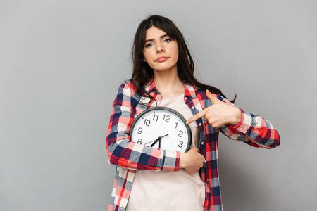 Image of displeased young lady standing isolated over grey background wall holding clock pointing.の写真素材