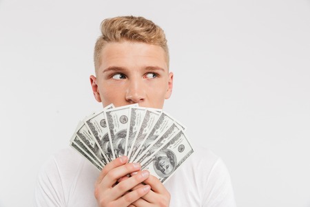 Portrait of a surprised teenage boy holding money banknotes at his face isolated over white backgroundの写真素材