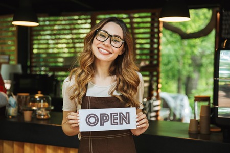Portrait of a pretty young barista girl in apron holding open sign board while standing at the cafeの写真素材