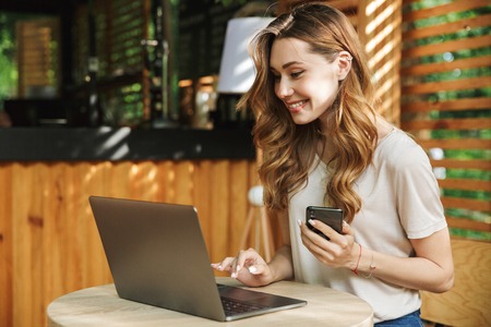 Portrait of a smiling young girl holding mobile phone while sitting with laptop computer at a cafe outdoorsの写真素材