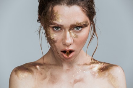 Beauty image of attractive brunette woman with soft healthy skin and golden makeup isolated over grey background looking camera holding pill in mouth.の写真素材