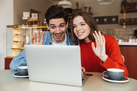 Cheerful young couple sitting at a cafe table together and waving hands at laptop computerの写真素材
