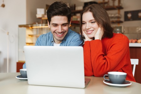 Cheerful young couple sitting at a cafe table together and looking at laptop computerの写真素材