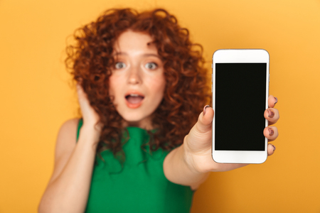 Portrait of a surprised redhead woman in dress showing blank screen mobile phone isolated over yellow backgroundの写真素材