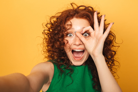 Close up portrait of a happy redhead woman in dress taking a selfie isolated over yellow backgroundの写真素材