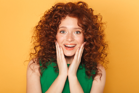Close up portrait of an excited curly redhead woman looking at camera isolated over yellow backgroundの写真素材