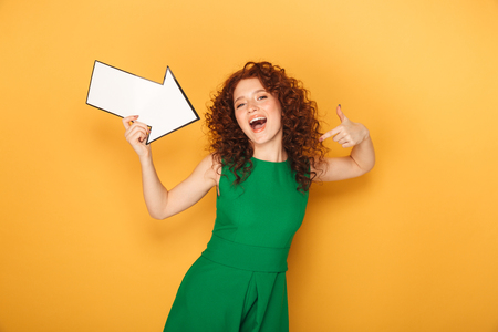 Portrait of a cheerful redhead woman in dress pointing away with an arrow isolated over yellow backgroundの写真素材